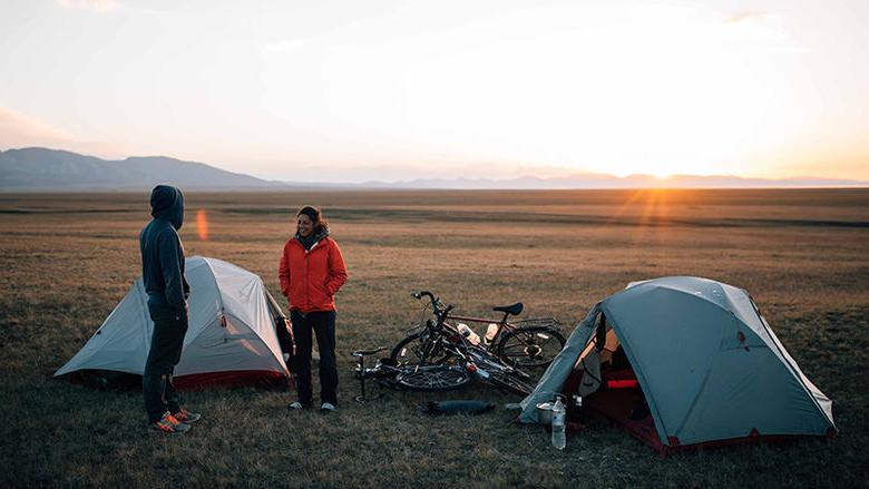 Nicole and a companion stand among their small white tents and a heap of bicycles on a grassy field, with the sun rising in the distance